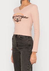 Hedvábně růžový dlouhý rukáv s žebrovaným vzorem a výstřihem do V, na kterém je grafický design s plameny a motocyklem v černé a oranžové barvě. Nosič má na sobě tmavé džíny.