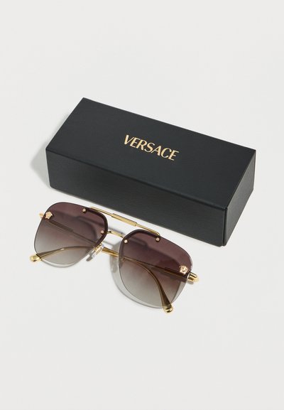 Μαύρα και χρυσά γυαλιά ηλίου Versace με φακούς μπράουν διαβάθμισης δίπλα σε μια μαύρη θήκη Versace με χρυσό λογότυπο σε λευκή επιφάνεια.