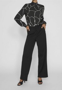 Černá dlouhá blůza s bílým abstraktním vzorem, vysokým límcem a nařasenými manžetami, kombinovaná s černými kalhotami s vysokým pasem a širokými nohavicemi, a sandály na otevřenou špičku.
