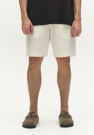 Mężczyzna ubrany w kremowe dzianinowe szorty, oliwkowozielone buty wsuwane i czarną koszulę, stojący na tle jednolitego białego tła.