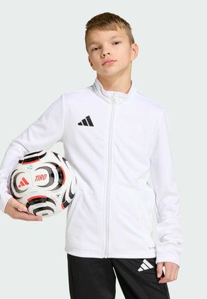 Chaqueta deportiva blanca con cremallera y cuello alto, con logo de tres rayas negras. Balón de fútbol negro con detalles en rojo sostenido en la mano izquierda.