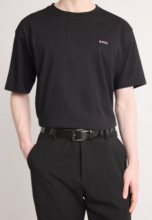 Persona con una camiseta negra de BOSS metida dentro de unos pantalones negros con un cinturón negro brillante, manos en los bolsillos, sobre un fondo liso y claro.