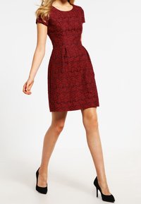 Femme marchant en robe rouge ajustée à manches courtes avec un motif floral, assortie à des chaussures à talons noirs, sur un fond blanc.