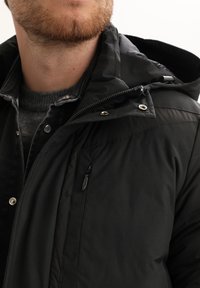 Giacca nera isolata con cappuccio, caratterizzata da accessori in metallo argentato, una tasca con zip e tessuto testurizzato. Indossata con un maglione grigio sotto.