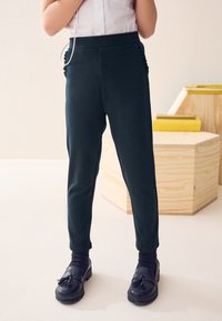 Pantaloni aderenti verde scuro realizzati in materiale morbido, con dettagli raggruppati ai lati e una cintura elastica, abbinati a mocassini neri e calzini.