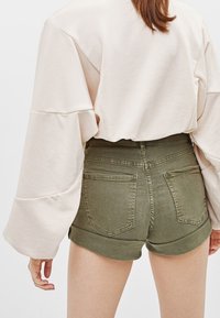 Beige sweatshirt med vida ärmar bärs över olivgröna jeansshorts med uppvikta kant, som har två bakfickor och en figurnära design.