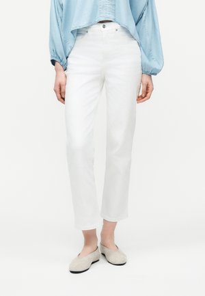 Naine, kes kannab valgeid sirge lõikega pükse, helerohelist lühikest pluusi ja beeže tasaseid kingi, seisab tavalise valge tausta ees.