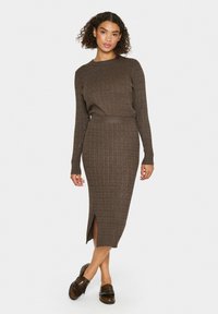 Robe en tricot côtelé marron avec manches longues et silhouette ajustée, dotée d'une fente latérale et d'un col arrondi. Portée avec des chaussures marron.