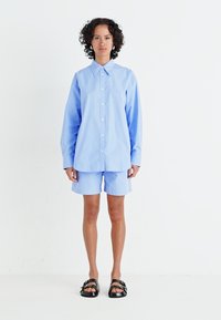 Cămașă button-up de culoare albastru deschis cu guler, asortată cu pantaloni scurți. Modelul poartă sandale negre. Material neted, croială lejeră.