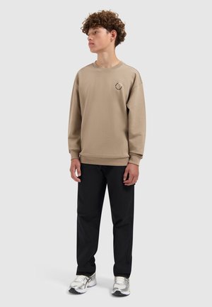 Beige katoenen sweatshirt met een ronde hals, geribbelde manchetten en een klein logo-detail. Gecombineerd met zwarte broeken en zilveren sneakers.