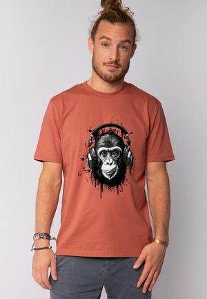 Mann mit Bart und Dutt, der ein rostfarbenes T-Shirt mit einer schwarz-weißen Grafik eines Gorillagesichts mit Kopfhörern trägt.