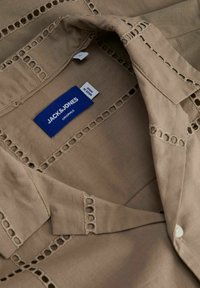Beige Baumwollhemd mit Kragen, das dekorative Ausschnittdetails entlang der Kanten aufweist. Enthält ein blaues Markenetikett mit der Aufschrift "JACK & JONES".