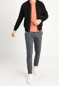 Černá bomber bunda přes korálové tričko, šedé úzké kalhoty a bílé tenisky. Bunda má zipový uzávěr a boční kapsy.