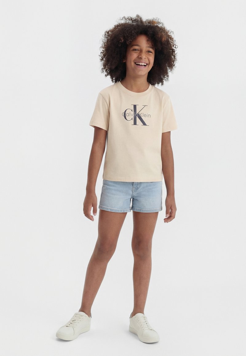 Niño sonriente con cabello rizado, vistiendo una camiseta beige de Calvin Klein, pantalones cortos vaqueros claros y zapatillas blancas, de pie frente a un fondo sencillo.