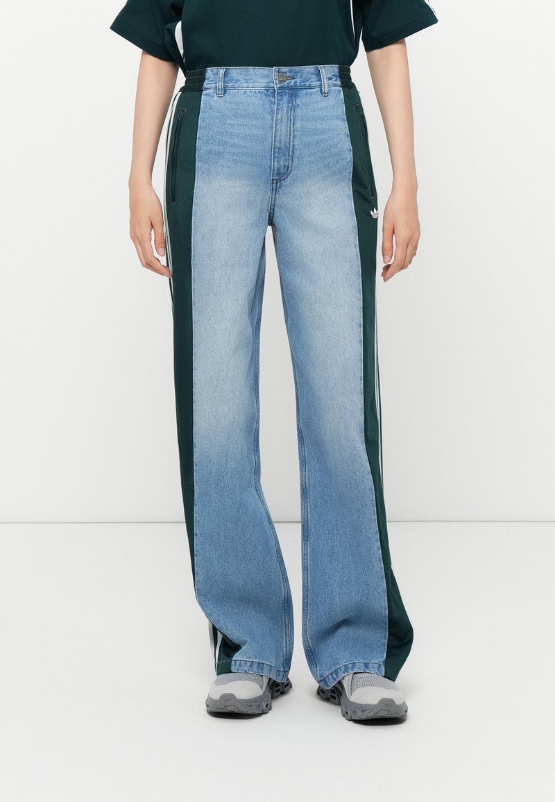 Persona che indossa pantaloni a gamba larga, con pannelli anteriori in denim blu e pannelli laterali in verde scuro, dotati di tasche con zip e strisce bianche.