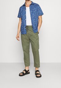 Man in een blauwe korte mouwen bloemetjes overhemd over een wit T-shirt, olijfgroene broek met trekkoord en zwarte sandalen, staand tegen een witte achtergrond.