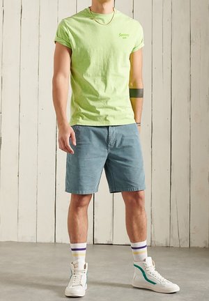 Man som bär en ljusgrön t-shirt med uppvikt ärm, blå shorts, vita höga sneakers och randiga strumpor, står mot en vit trävägg.