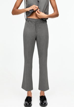 Femme portant un pantalon gris sur mesure et des mocassins noirs, boutonnant un gilet sans manches gris assorti, debout devant un fond blanc.