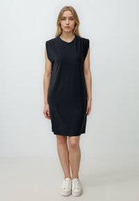 Schwarzes ärmelloses Kleid mit lockerer Passform, aus glattem Stoff gefertigt, mit Rundhalsausschnitt und knielangem Saum. Getragen mit weißen Sneakers.