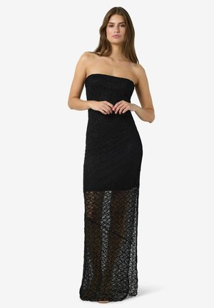 Nő, aki fekete, pánt nélküli csipkeruhát visel, szilárd miniszoknyával és átlátszó csipkével, amely a padlóig ér, fehér háttér előtt áll.