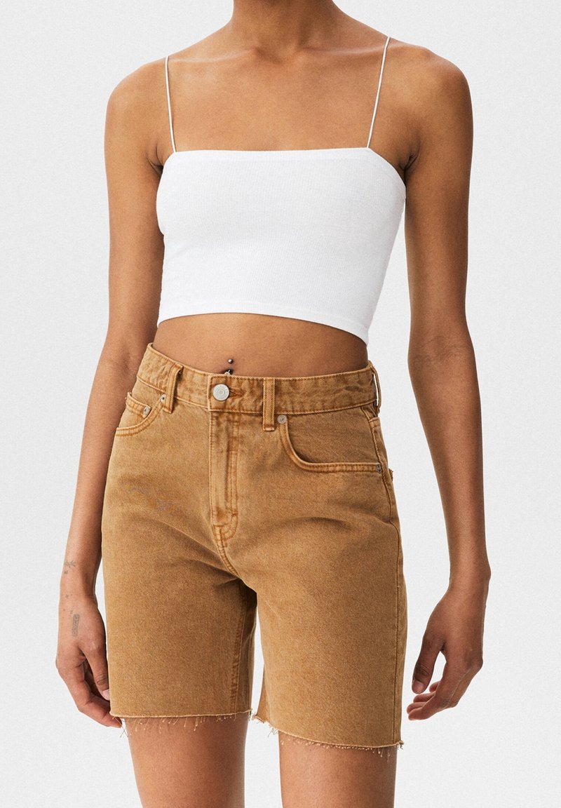 Femme portant un top court blanc à fines bretelles et un short en jean marron taille haute aux genoux avec des ourlets bruts, debout devant un fond uni.