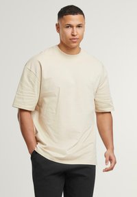 Beige Übergröße T-Shirt mit kurzen Ärmeln und Rundhalsausschnitt, aus weichem Stoff. Kombiniert mit schwarzen Hosen steht das Model mit den Händen in den Taschen.