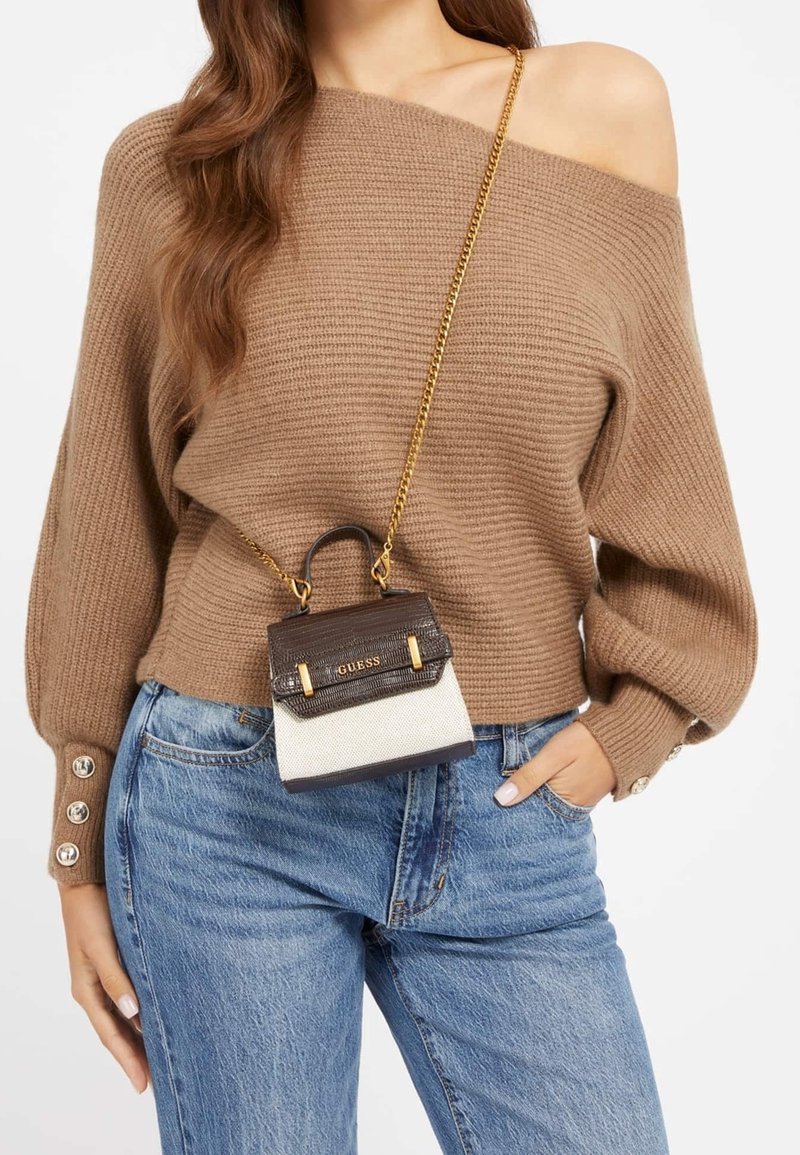 Femme en pull marron à épaules dénudées et jean bleu, portant un petit sac Guess marron et crème avec une bandoulière chaîne dorée croisée sur le corps.