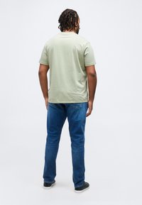 Άνδρας με πλεξούδες, που φοράει ανοιχτοπράσινη μπλούζα, μπλε τζιν και μαύρα αθλητικά παπούτσια, στέκεται με την πλάτη γυρισμένη σε λευκό ουδέτερο φόντο.