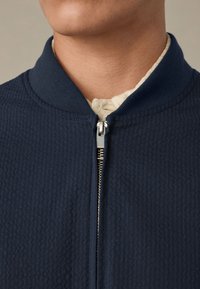 Artimo planas rodo asmens kaklą, dėvinčią tamsiai mėlyną tekstūruotą švarką su sidabriniu užtrauktuku, dalinai užsegtu virš kreminės spalvos marškinių.