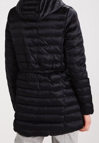 Svart puffjacka med huva, med kviltade sektioner och bälte i midjan. Slätt, glänsande material med en figurnära silhuett.