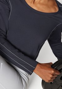 Marinefarbener, langärmliger Athletik-Oberteil mit grauen Nähten, getragen zu hellgrauen Sportleggings. Eine Hand hält eine schwarze Gewichtsscheibe.
