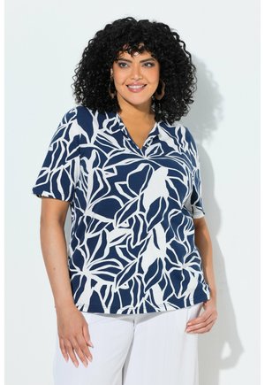 Uśmiechnięta kobieta o kręconych włosach, ubrana w granatowo-białą koszulę w liście i białe spodnie, stojąca na tle gładkiej jasnej ściany.