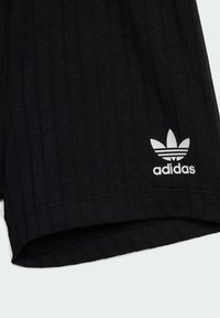 Tela negra acanalada con el logo de trébol de Adidas en blanco impreso cerca del borde inferior, mostrando detalles de la costura del dobladillo doblado.