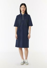 Marineblauwe jurk met korte mouwen, een kraag en een halflange rits, met twee borstzakken. Gladde textuur en knielange uitstraling.