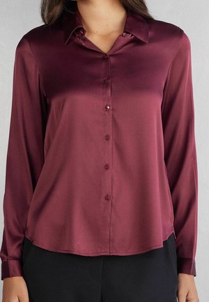 Kvinde iført en mørk bordeaux satinbluse med lange ærmer, knapper og krave samt sorte bukser, vist fra nakke til hofter.