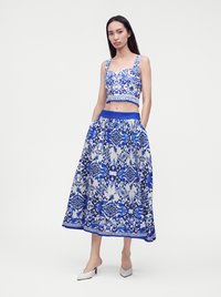 Γυναίκα που φοράει μπλε και λευκό crop top με σχέδια και μακριά φούστα με τσέπες, στεκόμενη σε λευκές ψηλοτάκουνες γόβες σε μονόχρωμο φόντο.
