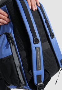 Mochila azul y negra con textura de malla acolchada, correas ajustables y una etiqueta de marca. Cuenta con un bolsillo con cremallera en el lado.