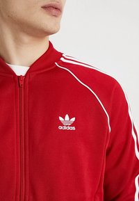 Punainen vetoketjullinen urheilutakki, joka on valmistettu pehmeästä kankaasta. Mukana valkoiset raidat hartioilla ja Adidaksen logo kirjailtuna rintaan.