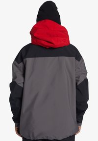Pánska bunda s červenou kapucňou, čiernou hornou časťou a šedou dolnou časťou. Má voľný strih a hladký, vodeodolný materiál.