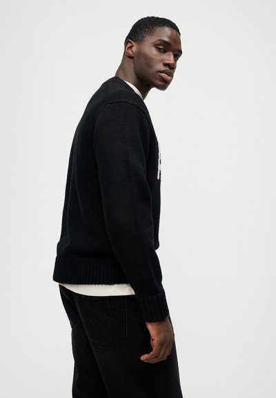 Fekete kötött pulóver, bordázott szegéllyel és mandzsettákkal, fehér ing felett viselve, minimális háttér előtt bemutatva.