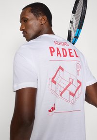 Biała bawełniana koszulka z czerwonym nadrukiem graficznym przedstawiającym plan kortu do padla. Na plecach nadruk z napisami "ADIDAS" i "PADEL".