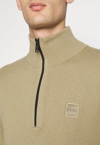 Khaki pulovr s čtvrtým zipem, vyrobený z žebrované látky, s černým zipem a malou čtvercovou nášivkou s logem na hrudi.