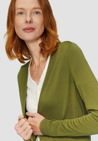 Groene cardigan van fijne breimateriaal, met een V-hals en lange mouwen, gedragen over een witte top met knopen aan de voorkant.