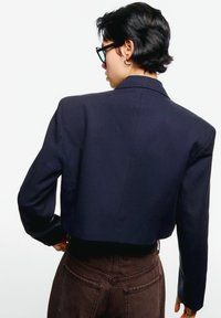 Short navy cropped blazer with padded shoulders, sharp lapels, and a smooth texture, paired with brown high-waisted pants.