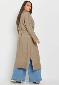 Beige stickad cardigan med krage, bältad i midjan, med ribbade manchetter och sidoflikar, bärs över ljusblå vida jeans.