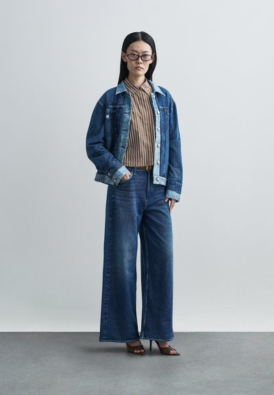 Casaco de ganga com gola azul-claro, camisa bege às riscas, jeans de pernas largas azuis e saltos abertos castanhos num fundo neutro.