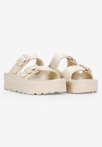 Sandales beiges avec une semelle épaisse et texturée, des brides larges et deux boucles ajustables. Fabriquées en matériau lisse et durable. Design ouvert à l'avant.