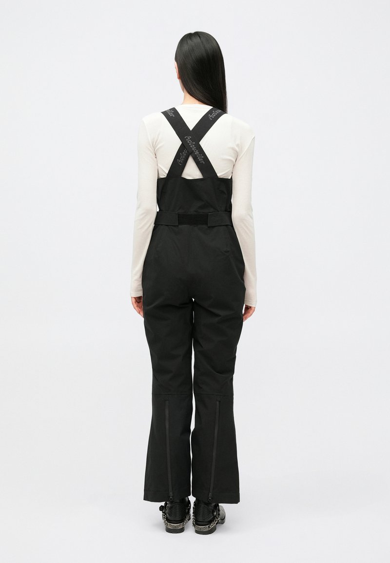 Black bib overalls with adjustable cross-back straps, tapered legs, zippers at the ankles, and a smooth fabric finish.