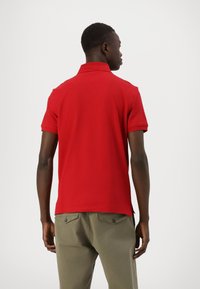 Camisa polo roja con cuello clásico, mangas cortas y dobladillo recto. Lleva puesta con pantalones verde olivo. Textura de tela suave.
