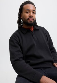 Sort pullover sweatshirt med krave og knapstolpe. Blødt stof og afslappet pasform. Båret over en mørkerød skjorte.